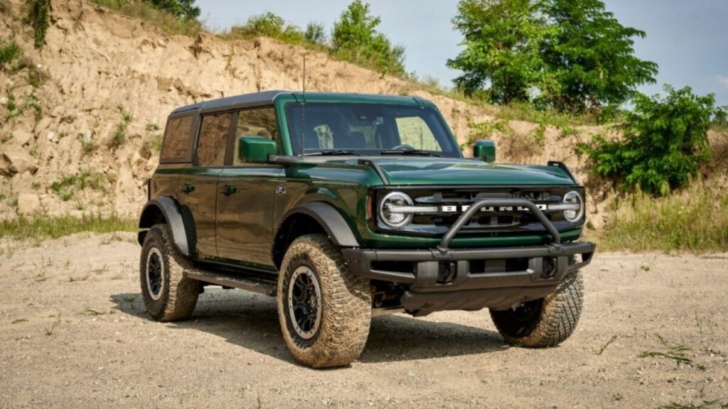 A forest green Ford Bronco SUV sits parked in front of a low desert cliff. The vehicles wheels are dirty but it is otherwise clean. We see it from a quarter angle. It is facing slightly to our right.