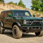 A forest green Ford Bronco SUV sits parked in front of a low desert cliff. The vehicles wheels are dirty but it is otherwise clean. We see it from a quarter angle. It is facing slightly to our right.